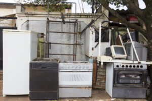 A row of old, discarded household appliances—including a refrigerator, dishwasher, stove, oven, and a computer—are lined up outdoors against a wall, beside a tree and some metal pipes.