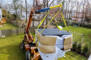 A construction forklift lifts a large, black rectangular object wrapped in cardboard and plastic over a grassy backyard, surrounded by trees and wooden fences.