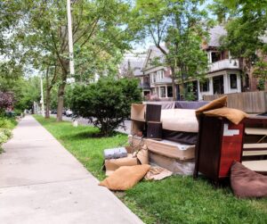 Old furniture and household items are piled on the grass next to a sidewalk in a residential neighborhood with large trees and houses in the background.