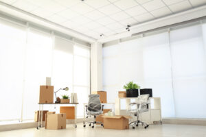 A bright office space with large windows, two chairs wrapped in plastic, several packed cardboard boxes, a desk lamp, plants, and white shelves, indicating an office move or setup.
