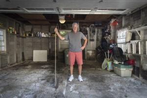 An older man wearing glasses, a gray shirt, red shorts, and white sneakers stands smiling and holding a broom in the middle of a worn, cluttered garage with various tools and storage items on the sides.