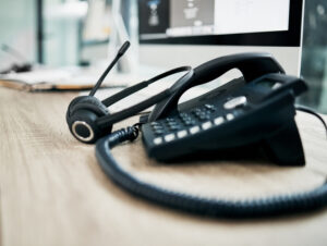 A black desk phone with a connected headset rests on a wooden office desk beside a computer monitor and open notebook, creating a typical workspace scene.